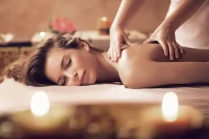 L’image montre une personne allongée, recevant un massage relaxant du dos dans une ambiance douce et chaleureuse. Des bougies floutées au premier plan renforcent l’atmosphère apaisante, tandis que la personne, les yeux fermés, semble totalement détendue et sereine